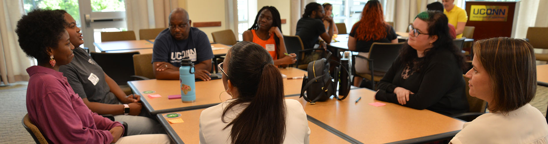 Five adult students and two professors sit at a table and talk.