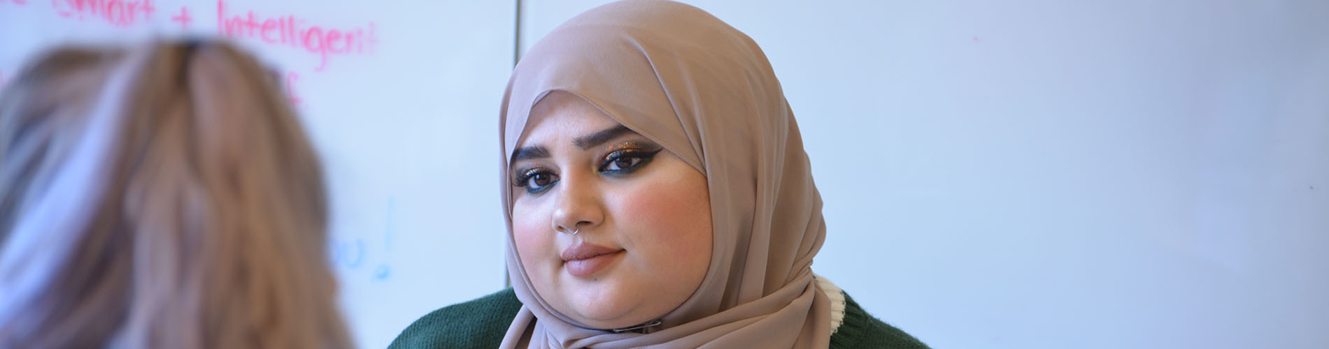 A graduate student wearing a headscarf smiles at a classmate.